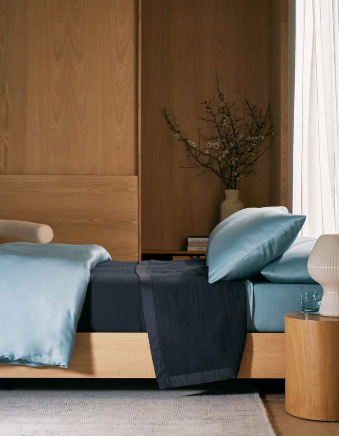 A modern bedroom features a light wood bed frame, blue satin bedding, and the Cozy Earth Waterfront Grid Matelasse Blanket in dark blue, with a wooden nightstand, ribbed white lamp, glass of water, and vase against wood-paneled walls and sheer curtains.|Color:Eclipse