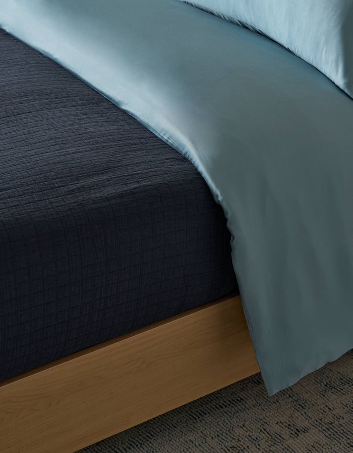 Close-up of a neatly made bed featuring the Cozy Earth Waterfront Grid Matelasse Blanket, a dark textured layer under a light blue satin comforter, on a wooden bed frame over a patterned carpet.|Color:Eclipse