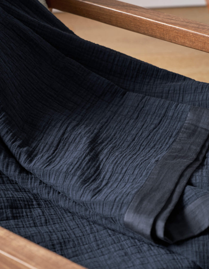 A close-up of Cozy Earth's Waterfront Grid Matelasse Blanket in dark navy, draped over wood, highlights its textured fabric with fine crinkles and a smooth edge.|Color:Eclipse