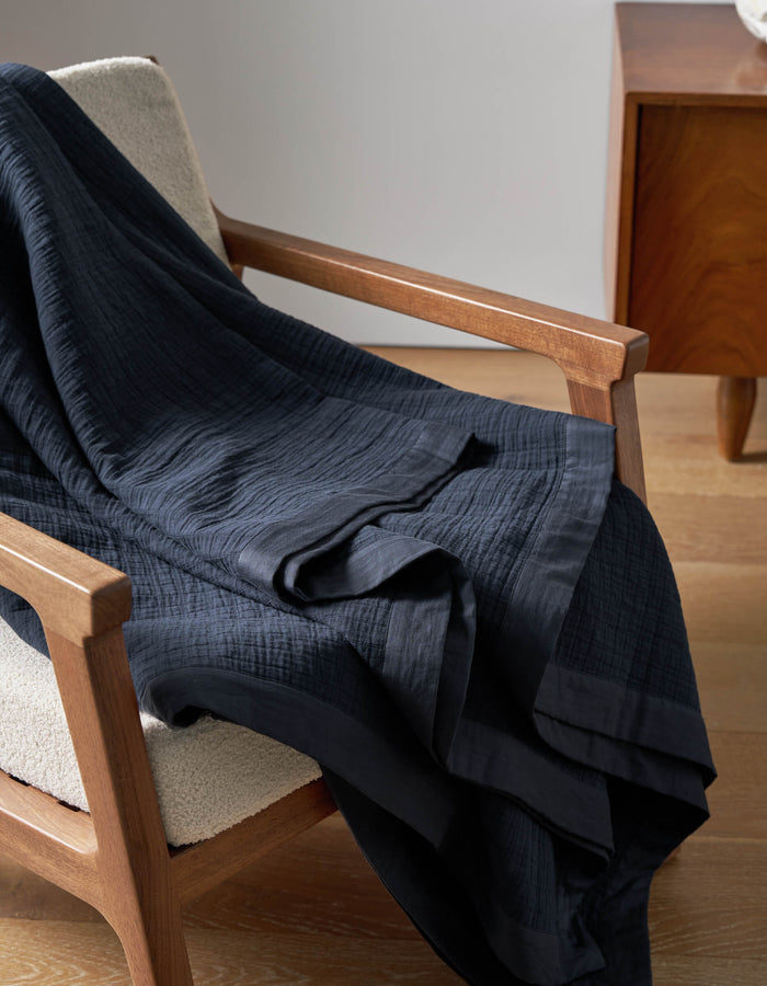 The Cozy Earth Waterfront Grid Matelasse Blanket in dark navy blue is draped over a light upholstered wooden armchair beside a wooden side table on a light wood floor.|Color:Eclipse
