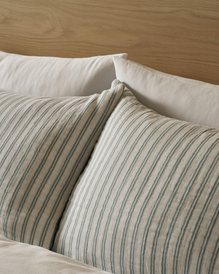 Two Cozy Earth Waterfront Shams, featuring light blue and white vertical stripes, rest against plain white pillows and a wooden headboard, creating a cozy and inviting bed setting.