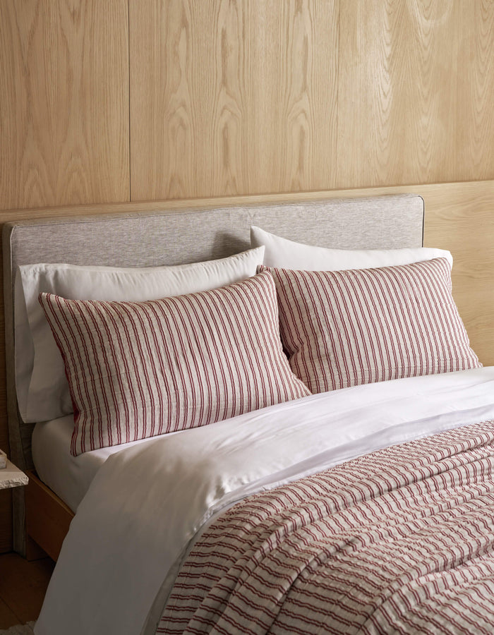 A Waterfront Coverlet Bundle by Cozy Earth, featuring white sheets, two pillows, and a comforter with red and white vertical stripes, is arranged on a bed with a light wood-paneled wall and a light gray upholstered headboard.|Color:Currant Stripe