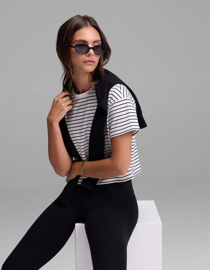 A woman wearing black sunglasses, black pants, and the Cozy Earth Women’s All Day Cropped Tee sits on a white block with a black sweater draped over her shoulders against a plain light gray background.|Color:Jet Black Stripe