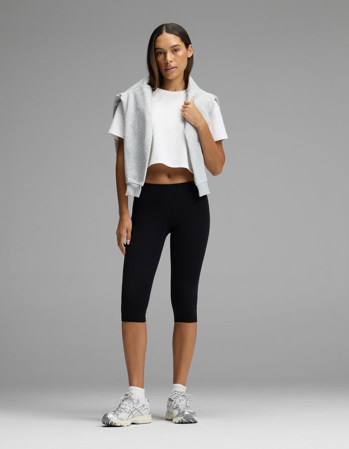A woman stands against a gray background, wearing the Women’s All Day Cropped Tee by Cozy Earth, paired with black leggings, white sneakers, and a gray sweatshirt draped over her shoulders. Her straight hair and relaxed, confident pose complete the look.|Color:White