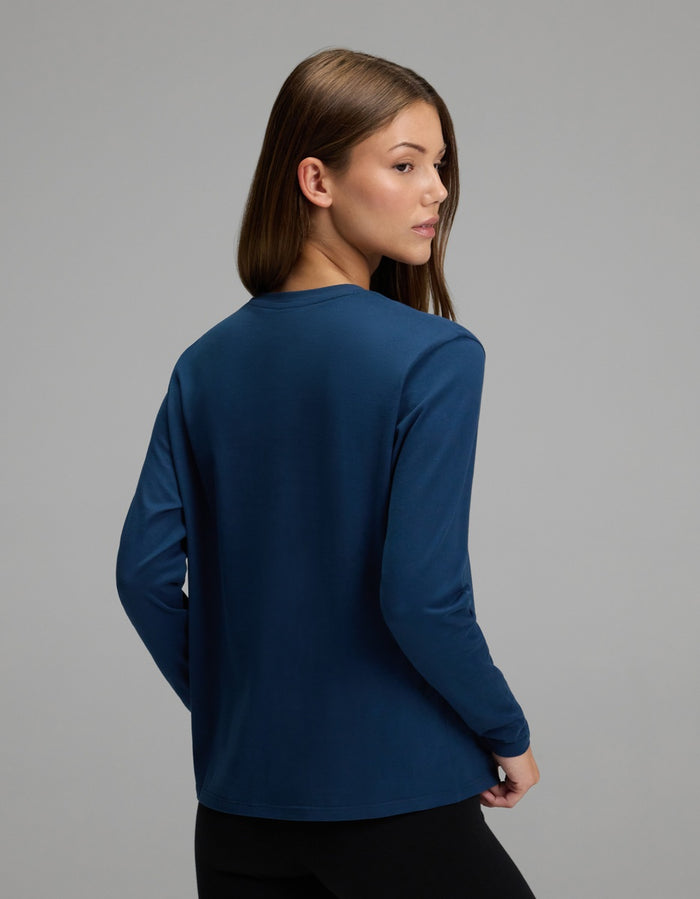 A woman with straight brown hair stands with her back to the camera and her head turned slightly over her left shoulder against a plain gray background, wearing the Cozy Earth Women’s All Day Long Sleeve Tee in dark blue paired with black pants.|Color:Obsidian