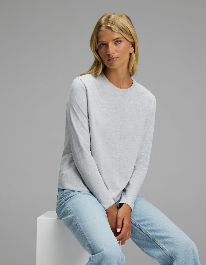 A woman with blonde hair sits on a white block against a plain gray background, wearing the Cozy Earth Women’s All Day Long Sleeve Tee in light gray and light blue jeans.|Color:Heather Grey