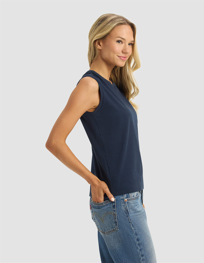 A woman with long blonde hair models the Cozy Earth Women's All Day Sleeveless Tee in navy, paired with blue jeans. She stands sideways with hands in pockets, smiling against a light gray background.|Color:Eclipse