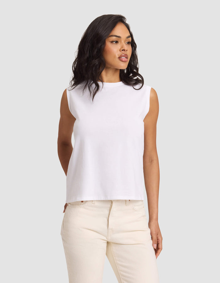 A woman with medium-length wavy dark hair wears the Cozy Earth Women's All Day Sleeveless Tee in white, paired with light beige pants, standing against a plain gray background and looking slightly to her left.|Color:White