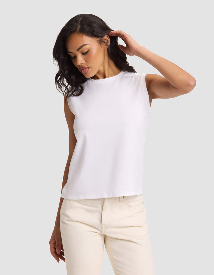 A woman models the Cozy Earth Women's All Day Sleeveless Tee in white, paired with light beige pants, standing against a plain light gray background and looking down with one hand touching her hair.|Color:White