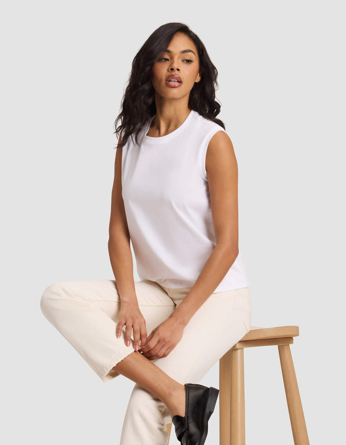 A woman with long, wavy dark hair sits on a wooden stool against a light gray background, wearing Cozy Earth's Women's All Day Sleeveless Tee in white, paired with cream pants and black shoes.|Color:White
