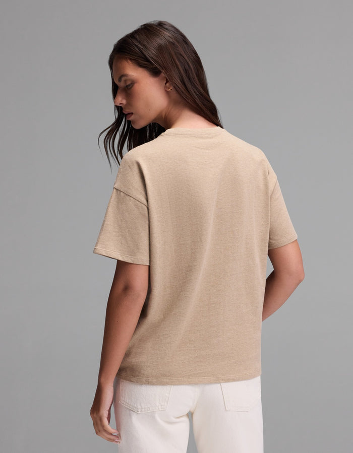 A woman with long brown hair stands facing away, wearing the Cozy Earth Women’s All Day Tee in beige and white pants against a plain gray background.|Color:Beech Heather