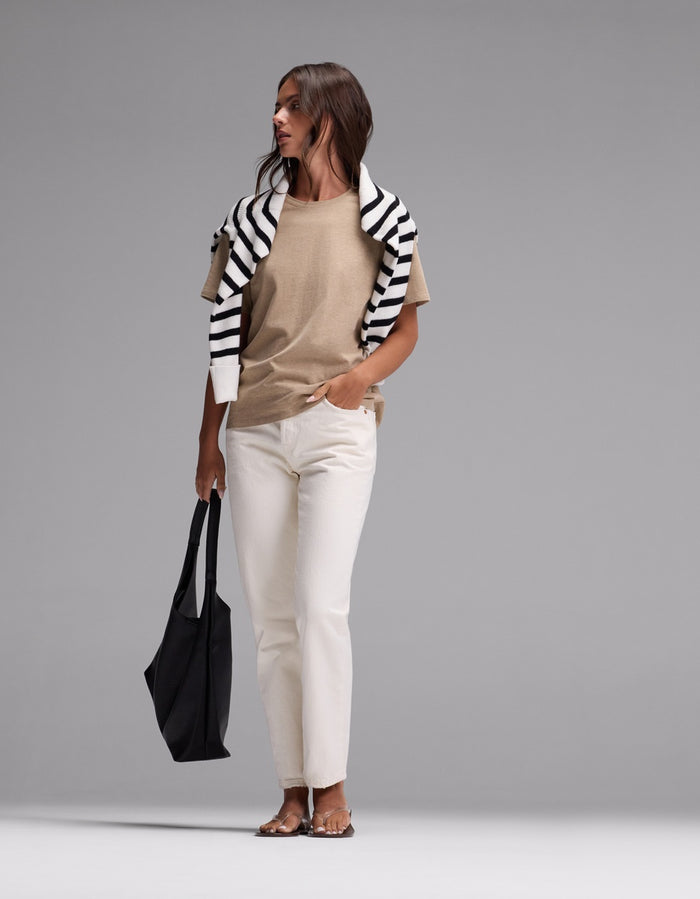 A woman stands against a plain gray background wearing Cozy Earth’s Women’s All Day Tee in beige, paired with white pants, sandals, and a black-and-white striped sweater draped over her shoulders, while holding a large black tote bag.|Color:Beech Heather