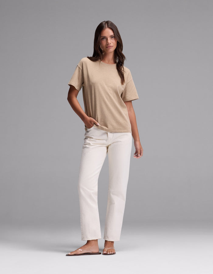 A woman stands against a plain gray background, wearing the Cozy Earth Women’s All Day Tee in beige, paired with white jeans and beige sandals. One hand is in her pocket as she looks slightly to the side.|Color:Beech Heather