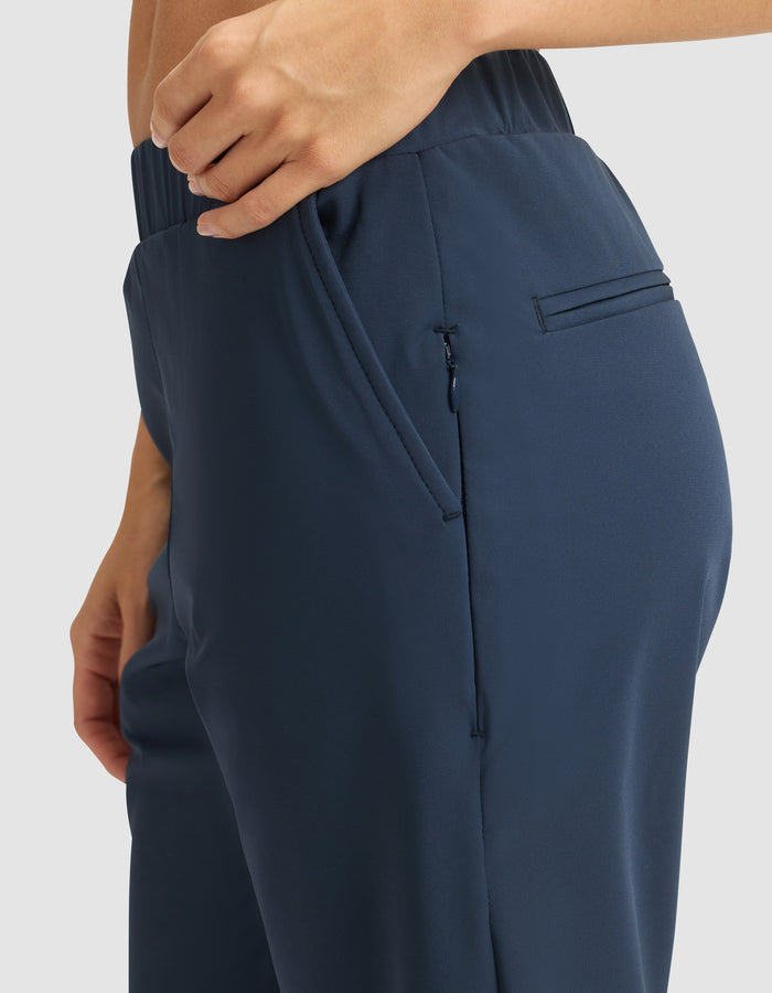A close-up of a person wearing Cozy Earth's Women's Always Pant in dark blue with an elastic waistband. One hand is near the zippered side pocket. The background is plain and light-colored. |Color:Eclipse