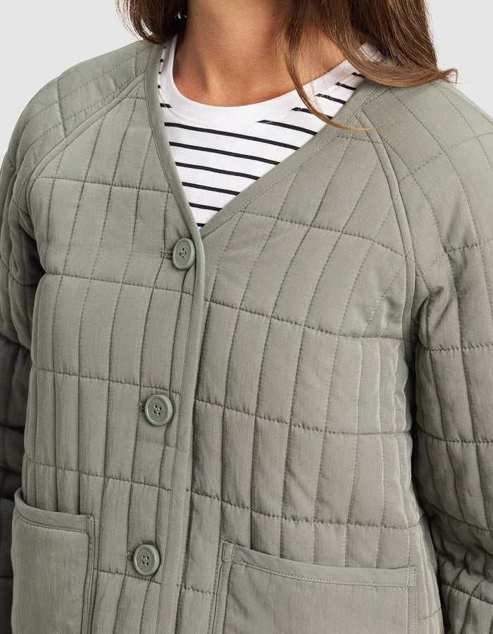 A woman wears the Cozy Earth Women's Andie Quilted Jacket in light gray with large buttons, a V-neck, and two front pockets over a white tee with black stripes. Only her upper body is visible.|Color:Cypress