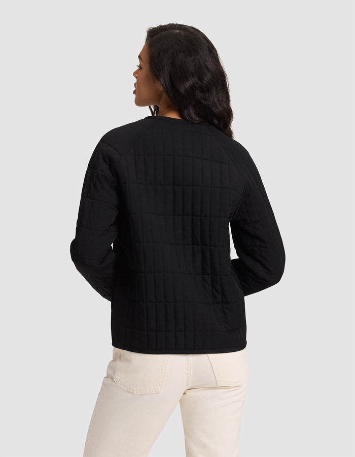 A woman is seen from the back wearing the Cozy Earth Women's Andie Quilted Jacket in black with light pants, standing against a plain light gray background.|Color:Jet Black