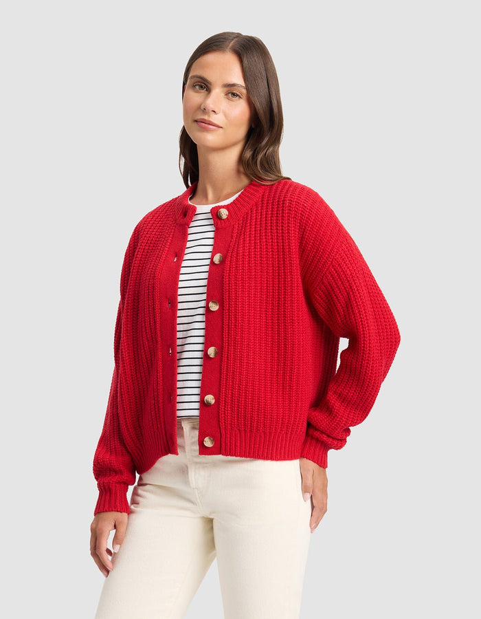 A woman with straight brown hair and a relaxed expression wears the Cozy Earth Women's Arden Cardigan in red over a white shirt with black stripes and cream-colored pants, standing against a plain background.|Color:Crimson