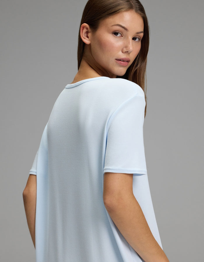 A young woman with light brown hair, wearing the Cozy Earth Women’s Bamboo Rib-Knit Boyfriend Sleep Dress in light blue, looks over her shoulder against a plain gray background.|Color:Frost