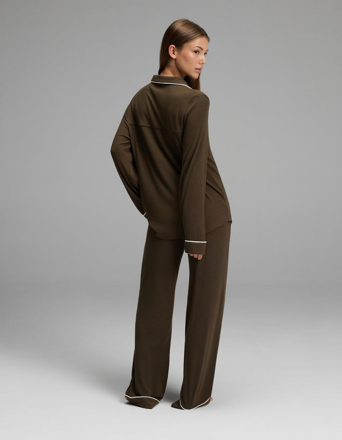 A woman stands in a studio, facing away but looking over her shoulder, wearing the Cozy Earth Women’s Bamboo Rib-Knit Classic Long Sleeve Pajama Set in a loose-fitting brown style with white trim. The background is plain and gray.|Color:Espresso