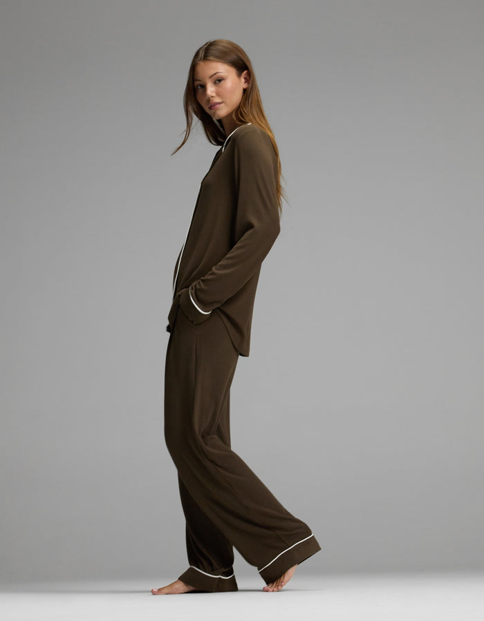 A woman stands barefoot in profile against a plain gray background, wearing the Cozy Earth Women’s Bamboo Rib-Knit Classic Long Sleeve Pajama Set, featuring a matching brown color with white piping on the collar, cuffs, and hems.|Color:Espresso