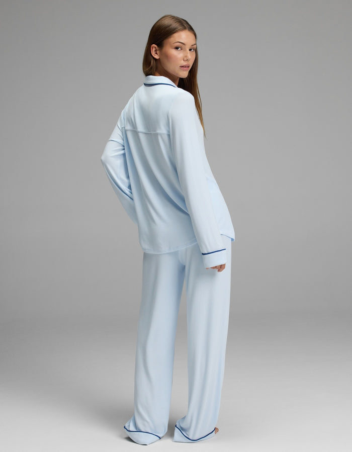 A woman models the Cozy Earth Women’s Bamboo Rib-Knit Classic Long Sleeve Pajama Set in light blue with dark trim, standing against a plain gray background and glancing over her shoulder.|Color:Frost