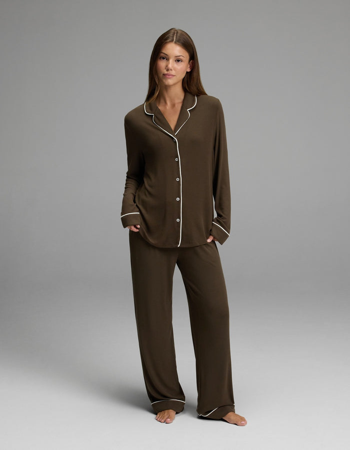 A woman stands barefoot on a gray background, looking at the camera with a neutral expression while wearing the Cozy Earth Women’s Bamboo Rib-Knit Classic Long Sleeve Pajama Top in dark brown with white trim.|Color:Espresso