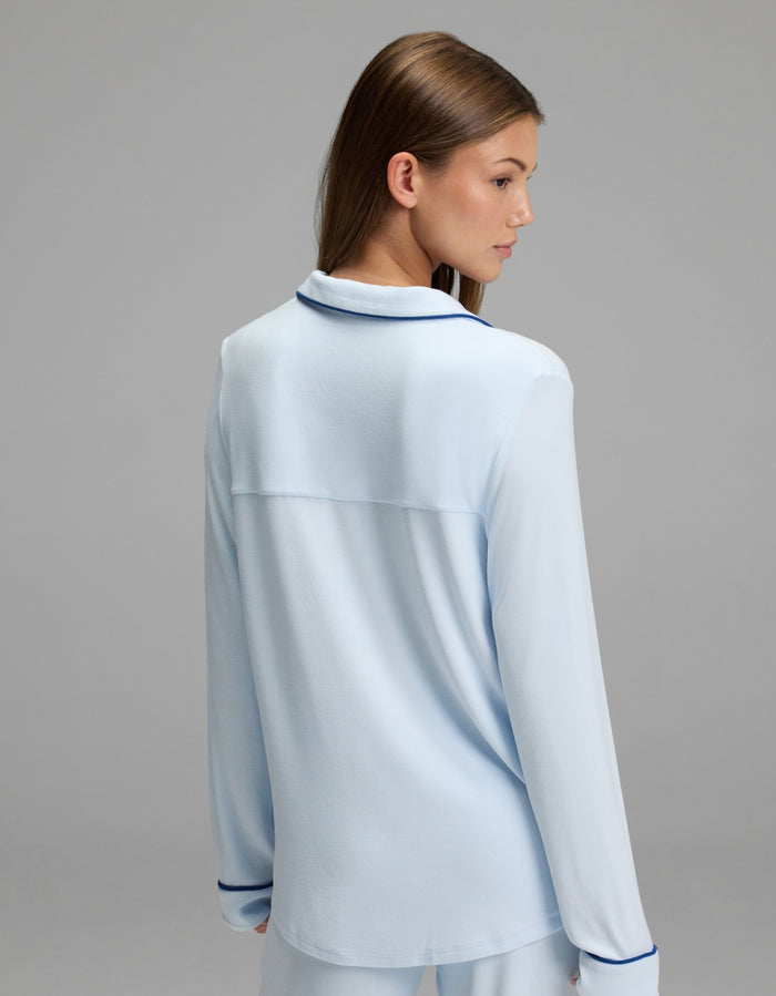 A woman with straight brown hair is standing and facing away, wearing the Cozy Earth Women’s Bamboo Rib-Knit Classic Long Sleeve Pajama Top in light blue with dark piping on the collar and cuffs, against a plain gray background.|Color:Frost