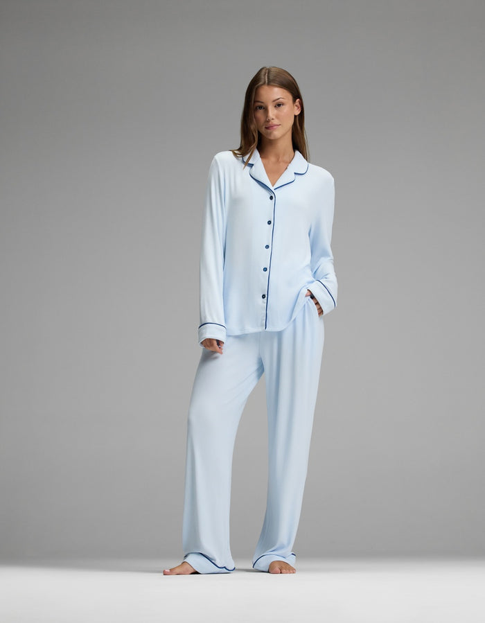 A woman stands barefoot against a plain gray background, looking relaxed with one hand in her pocket. She wears the Cozy Earth Women’s Bamboo Rib-Knit Classic Long Sleeve Pajama Top in light blue with black piping.|Color:Frost