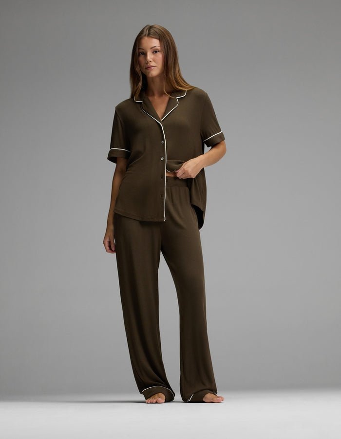 A woman stands barefoot against a plain gray background, wearing the Cozy Earth Women’s Bamboo Rib-Knit Classic Pajama Pant in brown with white trim, paired with a matching short-sleeve button-up top and loose pants.|Color:Espresso