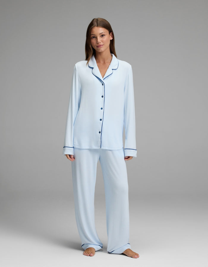 A woman stands barefoot against a plain gray background, wearing the Cozy Earth Women’s Bamboo Rib-Knit Classic Pajama Pant in light blue and a matching long-sleeve button-up pajama top with dark trim.|Color:Frost