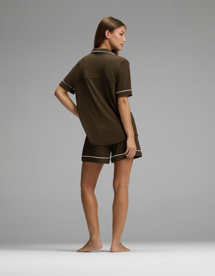 A person stands barefoot on a plain gray surface, facing away, wearing the Cozy Earth Women’s Bamboo Rib-Knit Classic Short Sleeve Pajama Set in dark brown with light trim.|Color:Espresso