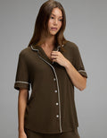 A woman with long brown hair wears the Cozy Earth Women’s Bamboo Rib-Knit Classic Short Sleeve Pajama Top, featuring dark brown fabric with white piping and buttons, while standing against a plain gray background.|Color:Espresso