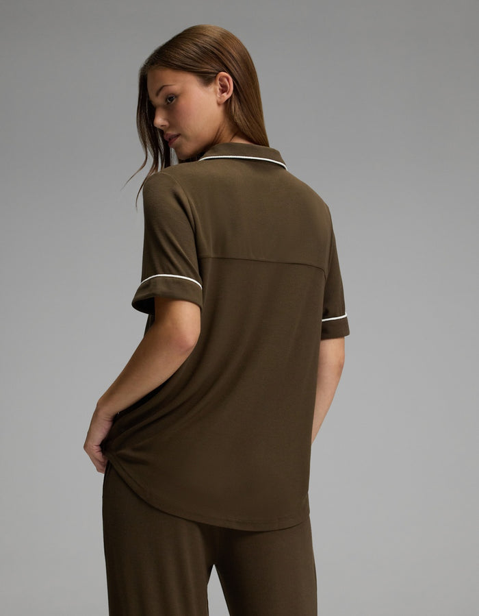 A woman stands facing away, wearing the Cozy Earth Women’s Bamboo Rib-Knit Classic Short Sleeve Pajama Top in brown with matching pants featuring white trim, set against a plain gray background.|Color:Espresso