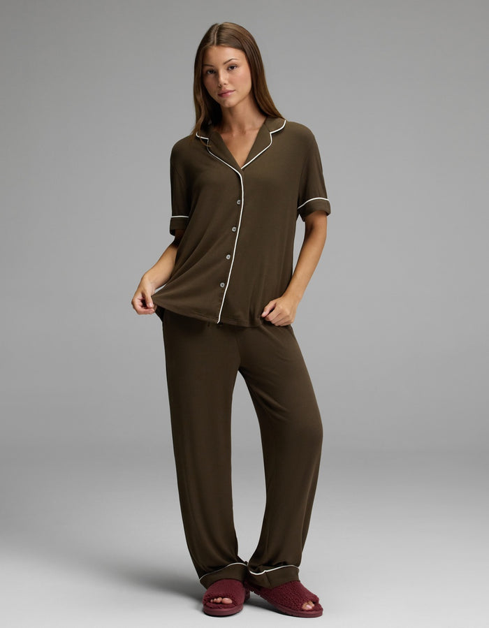 A woman stands against a plain grey background, holding the edge of her Cozy Earth Women’s Bamboo Rib-Knit Classic Short Sleeve Pajama Top in brown with white piping, and wears red slippers.|Color:Espresso