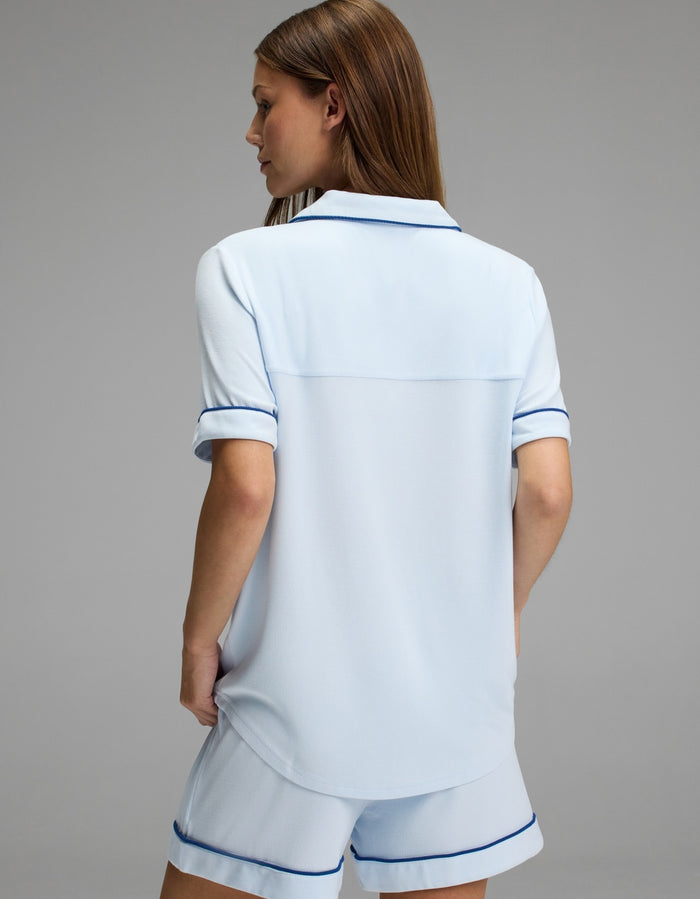 A person with long brown hair is shown from the back, wearing the Cozy Earth Women’s Bamboo Rib-Knit Classic Short Sleeve Pajama Top in light blue with dark blue trim, standing against a plain gray background.|Color:Frost