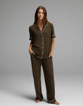 A woman stands barefoot, wearing the Cozy Earth Women’s Bamboo Rib-Knit Classic Short Sleeve & Pant Pajama Set in a loose-fitting, dark brown style with white piping. The set features a short-sleeve, button-up top and long pants. She is posed against a plain gray background.|Color:Espresso