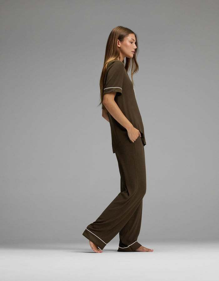 A woman with long hair stands barefoot in profile, wearing the Cozy Earth Women’s Bamboo Rib-Knit Classic Short Sleeve & Pant Pajama Set—a loose-fitting brown ensemble with white trim—against a plain gray background.|Color:Espresso