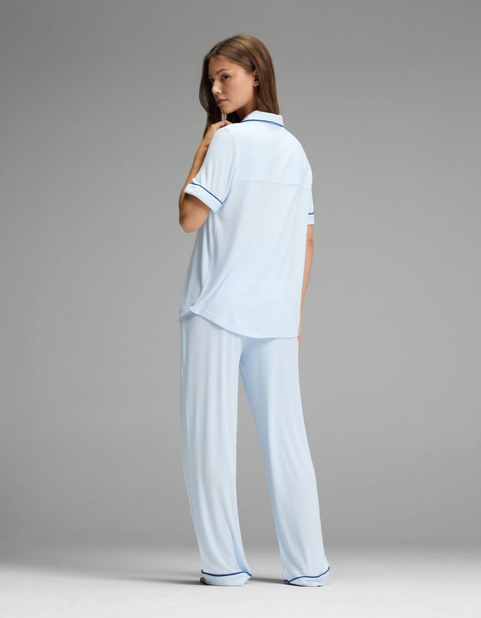 A woman stands against a plain gray background, looking over her shoulder and wearing the Cozy Earth Women’s Bamboo Rib-Knit Classic Short Sleeve & Pant Pajama Set in light blue with dark trim.|Color:Frost