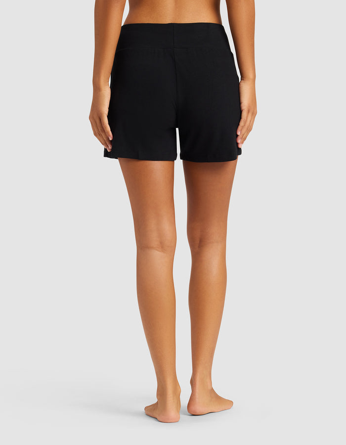 A person stands barefoot with their back to the camera, wearing Cozy Earth Women’s Bamboo Rib-Knit Lounge Shorts in black, no top, against a plain light gray background.|Color:Black