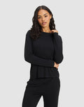 A woman with wavy dark hair models the Cozy Earth Women's Bamboo Stretch-Knit Long Sleeve Lounge Tee in black, standing against a plain light gray background with one hand on her shoulder and the other crossing her waist. |Color:Black