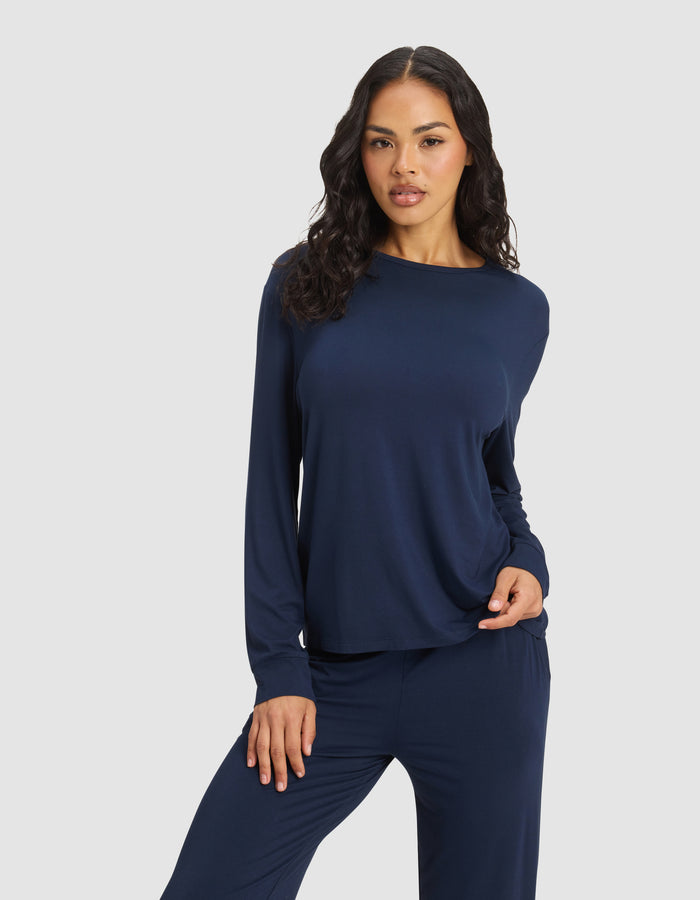 A woman with long, wavy dark hair models the Cozy Earth Women's Bamboo Stretch-Knit Long Sleeve Lounge Tee in navy blue, paired with matching pants, standing before a light gray background with one hand on her hip.|Color:Navy