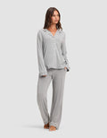 A woman stands barefoot against a plain light background, wearing the Cozy Earth Women's Bamboo Stretch-Knit Long Sleeve Pajama Set in light grey with white piping, featuring a button-up top and matching pants.  |Color:Grey