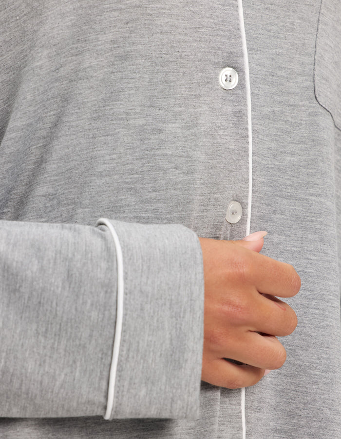 A close-up shows a person wearing the Cozy Earth Women's Bamboo Stretch-Knit Long Sleeve Pajama Top, gray with white piping, their hand resting near the chest area.|Color:Grey