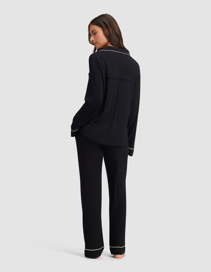 A woman stands barefoot, turned slightly away, wearing the Cozy Earth Women's Bamboo Stretch-Knit Long Sleeve Pajama Set in black with white piping, against a plain light gray background. |Color:Black