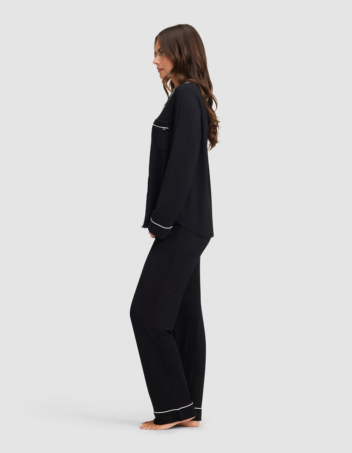A woman stands barefoot in profile against a light gray background, wearing the Cozy Earth Women's Bamboo Stretch-Knit Long Sleeve Pajama Set in black with white trim. |Color:Black