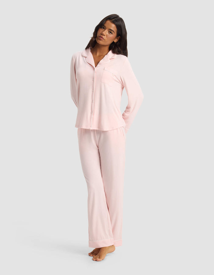 A woman with long dark hair stands barefoot against a light gray background, wearing Cozy Earth’s Women's Bamboo Stretch-Knit Long Sleeve Pajama Set—light pink, with a button-up top and matching pants. Her arms are relaxed behind her back.|Color:Blossom