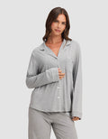 A woman with long brown hair wears the Cozy Earth Women's Bamboo Stretch-Knit Long Sleeve Pajama Top in light gray with white piping, standing against a plain background. She holds the shirt closed with one hand and smiles slightly.|Color:Grey