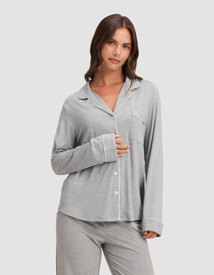 A woman with long brown hair wears the Cozy Earth Women's Bamboo Stretch-Knit Long Sleeve Pajama Top in light gray with white piping, standing against a plain background. She holds the shirt closed with one hand and smiles slightly.|Color:Grey