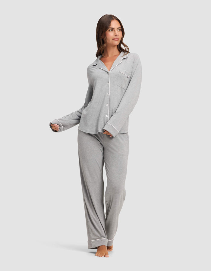A woman stands barefoot, looking to the side, wearing the Cozy Earth Women's Bamboo Stretch-Knit Long Sleeve Pajama Top in light gray with white trim and a chest pocket, paired with matching loose pants. One hand is slightly raised.|Color:Grey