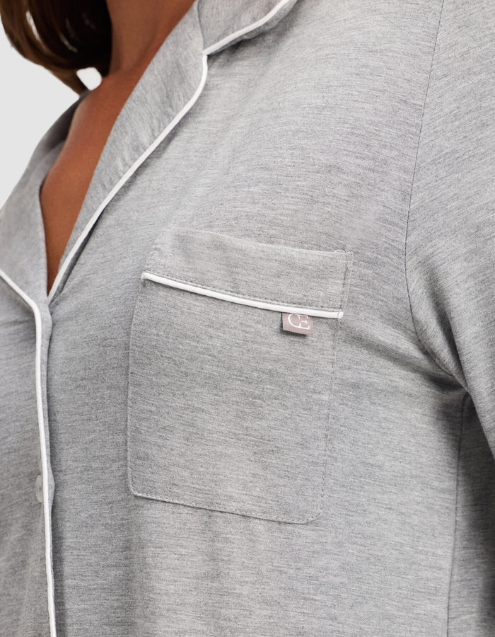 Close-up of a woman wearing Cozy Earth's Bamboo Stretch-Knit Long Sleeve Pajama Top in light grey, featuring white piping on the collar, pocket, and button line. A small fabric tag accents the chest pocket.|Color:Grey
